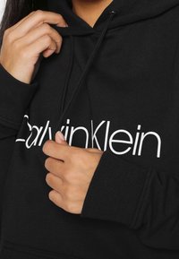 Felpa nera in un misto cotone, con il logo bianco "Calvin Klein". Include una tasca frontale e un cappuccio regolabile con coulisse.