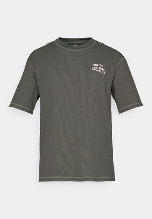 Kortermet t-skjorte i mørkegrønn, teksturert stoff med ribbestruktur. Har et hvitt "KARMA"-motiv på brystet og kontrasterende sømmer.