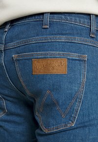 Siniset farkut, joissa on ruskea nahkapaikka ja "Wrangler"-logo, erottuva ommel ja ainutlaatuinen "W"-muotoinen takataskusuunnittelu.