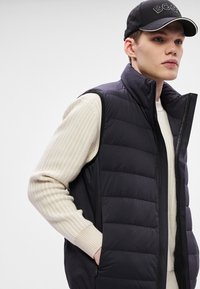 Polsterēta melna veste ar augstu apkakli, rāvējslēdzēja aizdari un sānu kabatām, saskaņota ar teksturētu krēmkrāsas džemperi un melnu cepuri ar baltām detaļām.