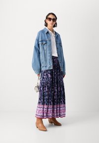 Veste en denim, blouse blanche, jupe longue fluide avec des motifs floraux bleu marine et rose, bottes de cheville beige et lunettes de soleil marron.