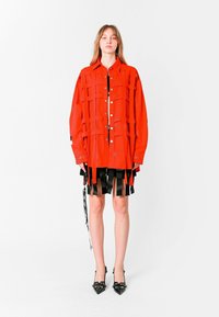 Camisa roja de gran tamaño con paneles estructurados, flecos negros en el dobladillo y botones brillantes. Combina con una falda negra y tacones negros de punta.