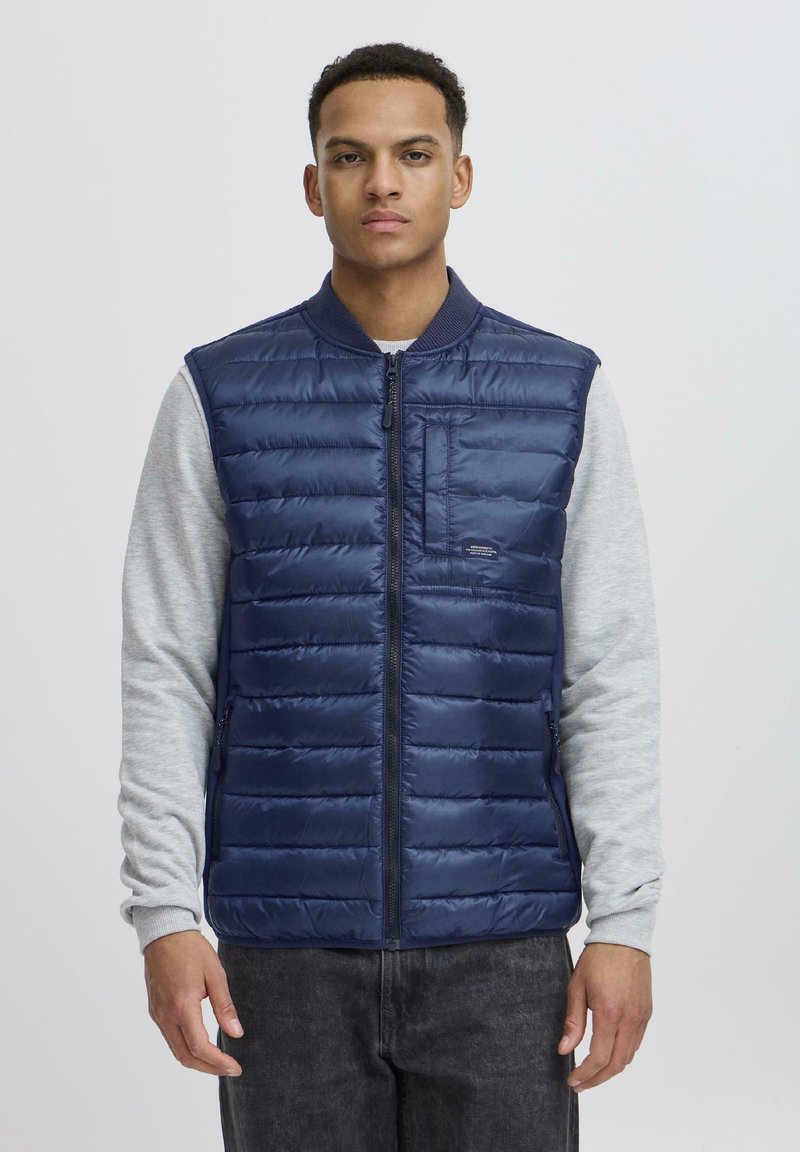 Muškarac u tamnoplavom sako od jastučića preko svijetlosive košulje s dugim rukavima i tamnim trapericama, stoji naslonjen uz jednostavnu svijetlosivu pozadinu.