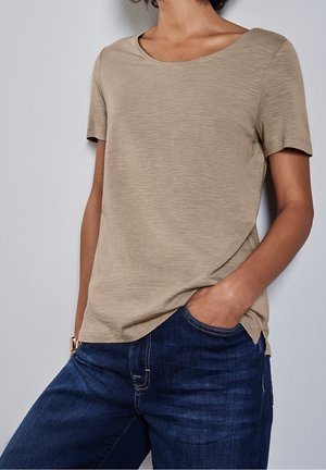 Person iført en beige kortærmet t-shirt og mørkeblå jeans med den ene hånd i lommen mod en lys grå baggrund.