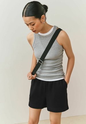 Noor naine, kellel on mustad juuksed kuklas, kannab halli varrukateta topi, musti šorte ja musta ristihoidsa, vaadates alla.