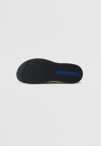Juodas gumos padas su tekstūruota raštu, su užrašu "BIRKENSTOCK" mėlyna spalva. Viršus yra smėlio spalvos su lygaus paviršiaus apdaila.