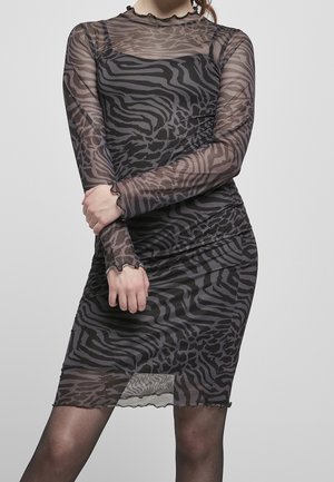 Kobieta w przezroczystej, czarno-szarej sukience z nadrukiem zwierzęcym, z długimi rękawami i falbanami na krawędziach, ze skrzyżowanymi ramionami, pozująca na jednolitym szarym tle.