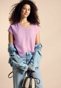 Blusa lilac a maniche corte, giacca di jeans leggera, jeans blu a vita alta e una piccola borsa dalla forma arrotondata nei toni neutri.