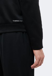 Gros plan de la main droite d'une personne à côté d'une veste et d'un pantalon noirs Therma-FIT avec poignets et taille élastiques.