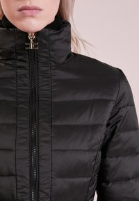 Czarna kurtka puchowa z wysokim kołnierzem, pikowaną fakturą i centralnym zamkiem błyskawicznym z złotymi elementami. Gładki, lekki materiał.