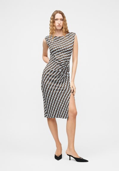 Γυναίκα με κυματιστά μαλλιά που φοράει ένα αμάνικο φόρεμα με μοτίβο μέχρι το γόνατο με πλευρική σχισμή, συνδυασμένο με μαύρες γόβες μυτερές.