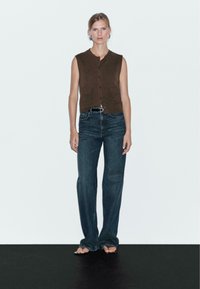 Gilet sans manches marron avec des boutons à l'avant et deux poches, associé à un jean en denim bleu foncé légèrement évasé. Fond neutre.