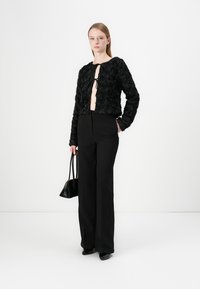 Veste noire texturée, courte, avec un motif floral, ouverte à l'avant et fermée par un lien ; associée à un pantalon noir taille haute et un petit sac à main.