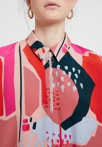 Blouse colorée en rose, rouge et marine avec des motifs géométriques. Dotée d'un col à revers et d'une fermeture éclair frontale avec une tirette circulaire.