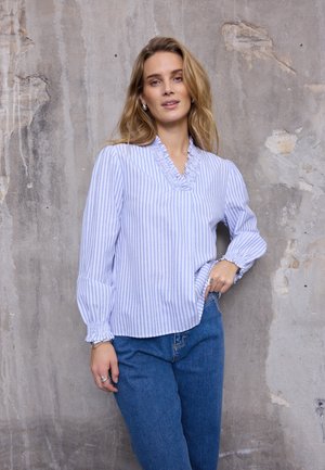 Blauw-wit gestreepte blouse met ruches bij de halslijn en elastische manchetten, gecombineerd met een blauwe high-waist jeans. Gestructureerde grijze achtergrond.