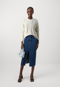 Strickpullover in Creme mit einem strukturierten Muster, kombiniert mit einem blauen Jeans-Midirock mit einem vorderen Schlitz. Hält eine kleine hellblaue Handtasche.