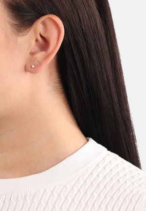 Pendiente de oro, de forma redonda y pequeña, con superficie brillante, llevado en la oreja izquierda. Cabello oscuro y liso parcialmente visible, suéter de textura ligera.