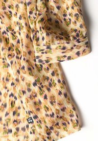 Blusa ligeramente transparente en tono beige claro con un patrón de estampado de leopardo multicolor. Presenta puños enrollados y una pequeña etiqueta de logo negra.