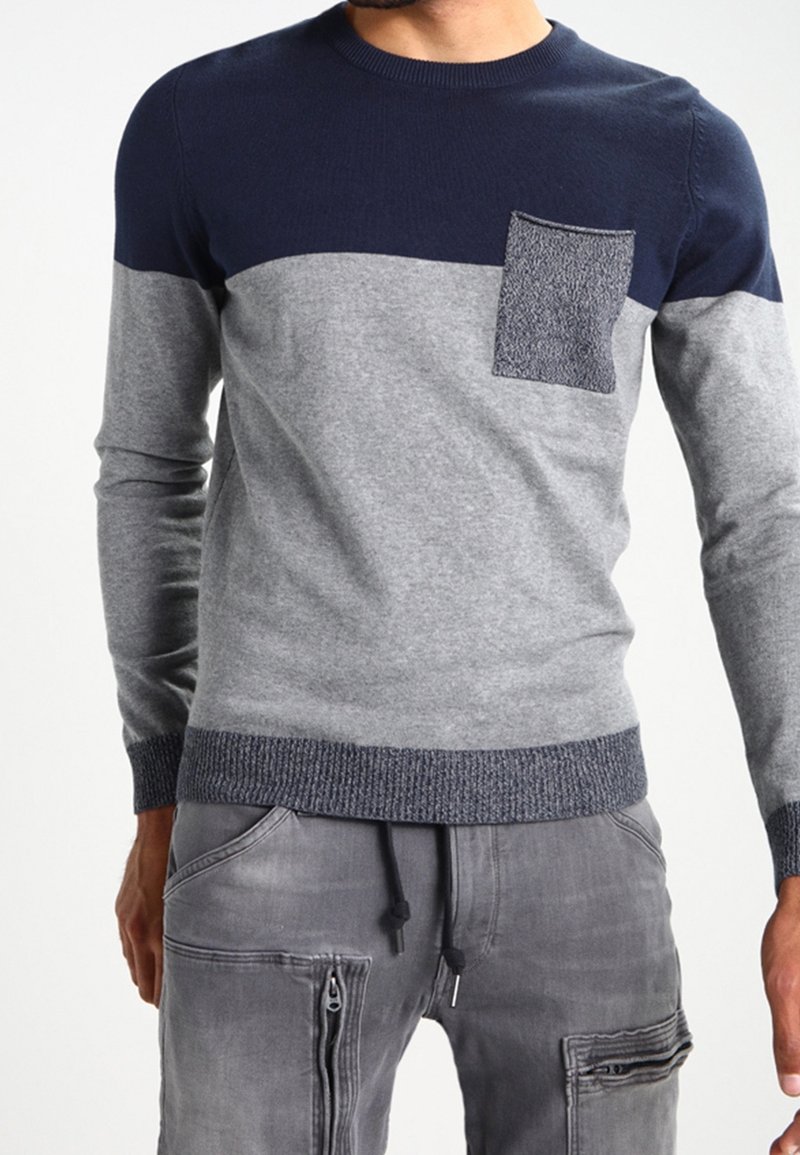 Homme portant un pull à manches longues bicolore bleu marine et gris avec une poche poitrine texturée, associé à un pantalon cargo gris avec des fermetures éclair.