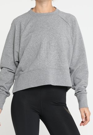 Personne portant un sweat-shirt gris ample à manches longues et un legging noir taille haute sur un fond uni.
