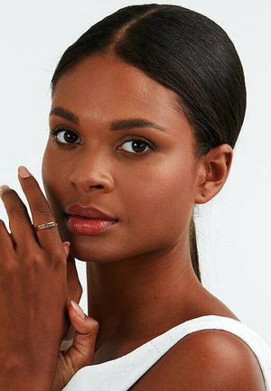 Goldring mit einem gedrehten Design, der an einem Finger getragen wird, mit einem glatten, warmen Teint und dunkelbraunem, nach hinten gestyltem Haar. Neutraler Hintergrund.