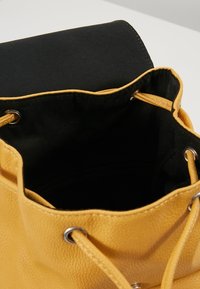 Hořčicově žlutá kožená taška na stahovací šňůrku s černou textilní podšívkou a stříbrnými kovovými oky na bílé ploše.