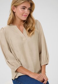 Blouse beige à manches longues avec un décolleté en V, des manches froncées et une texture douce. Associée à un jean bleu, mettant en avant une coupe décontractée.