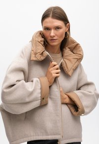 Bež jakna od vune s mješavinom s odvojivim smeškim pufastim ovratnikom, oversized siluetom, zatvaračem sprijeda i okruglim rukavima s gumbima.