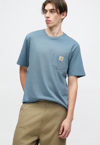 Světle modré bavlněné tričko s kulatým výstřihem, krátkými rukávy a kapsou na levé straně hrudi, která obsahuje nášivku s logem. Jednoduchá barva, uvolněný střih.