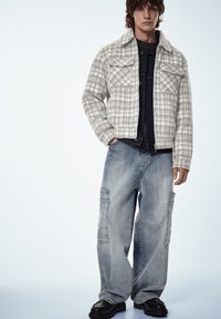 Vaaleanharmaa ruudullinen takki, jossa kaksi rintataskua, tumman farkkupaidan päällä. Löysät siniset farkut, joissa on taskuja, mustat korolliset kengät.