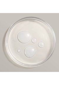 Klar petriskål med fyra droppar av genomskinlig vätska i olika storlekar, som vilar på en slät beigefärgad yta.