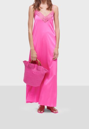 Vrouw draagt een felroze satijnen maxi-jurk met kanten halslijn, houdt een gestructureerde roze handtas met bamboe handvatten vast en draagt roze sandalen.