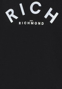 T-shirt in cotone nero con il carattere bianco in grande che recita "RICH" sopra "john RICHMOND." Il testo è rifinito con una superficie lucida ed è centrato sulla parte anteriore.