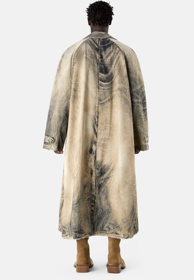 Homme portant un long manteau beige surdimensionné avec des motifs abstraits noirs et des bottines en daim couleur fauve, vu de dos.