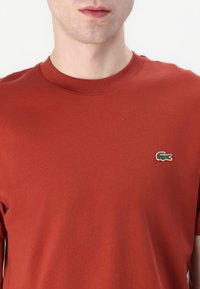 Tricou roșu din bumbac, cu guler rotund, mâneci scurte și un mic logo de crocodil verde brodat pe pieptul stâng.
