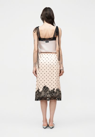 Γυναίκα που φοράει κρεμ σατέν φούστα με μαύρες πουά και δαντελένιο τελείωμα, σε συνδυασμό με αμάνικο τοπ με διάφανα μαύρα κορδόνια στους ώμους, γυρισμένη προς τα πίσω.