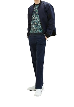 Navy bomber jacket over a green floral pattern shirt, paired with navy trousers and white sneakers. Simple, fitted design, and smooth textures.