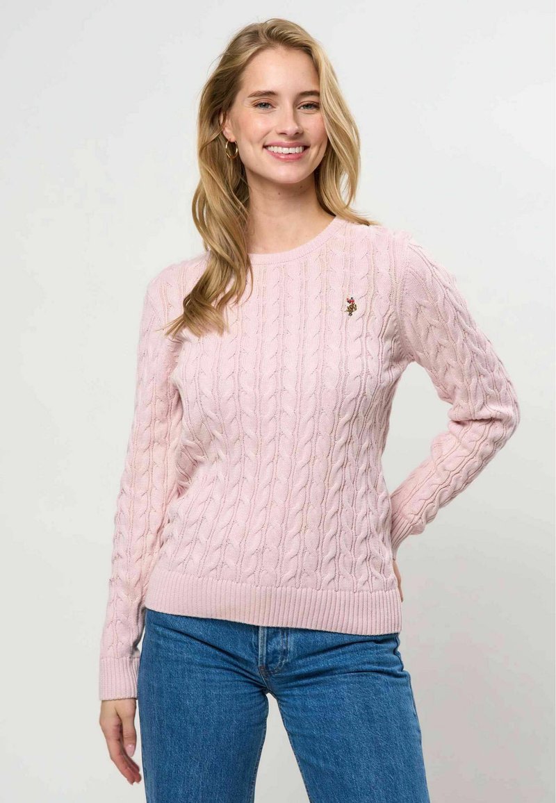 Femme souriante aux longs cheveux blonds porte un pull torsadé rose et un jean bleu, debout avec une main sur la hanche.