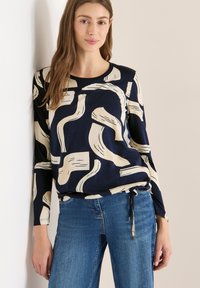 Nuori nainen pukeutuneena tummansiniseen pitkähihaiseen paitaan, jossa on abstrakteja, beigejä siveltimen vetoja, yhdistettynä sinisiin farkkuihin, seisoo vaaleaa seinää vasten.