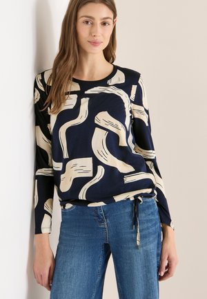 Młoda kobieta w granatowej koszuli z długim rękawem zdobionej abstrakcyjnymi beżowymi pociągnięciami pędzla, w niebieskich dżinsach, stojąca na tle jasnej ściany.