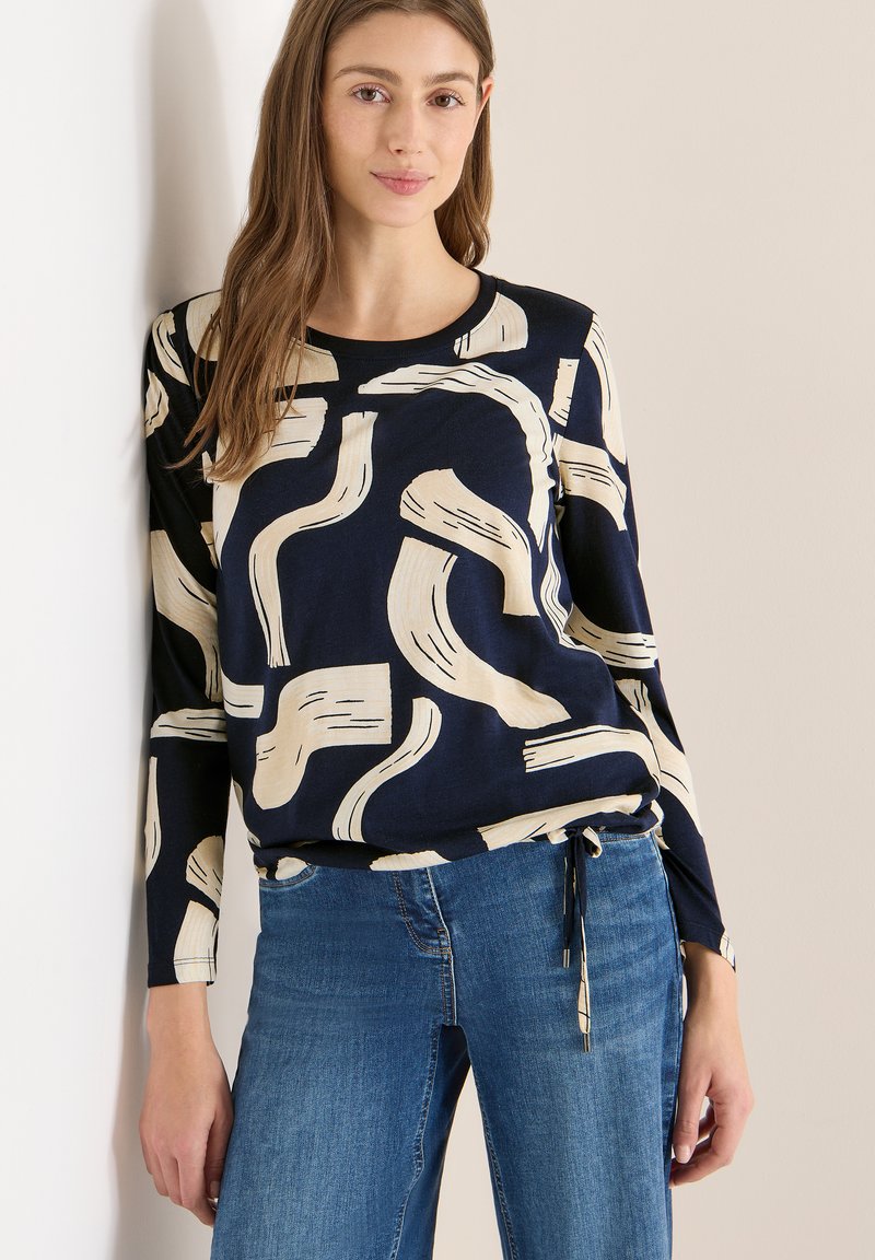 Nuori nainen pukeutuneena tummansiniseen pitkähihaiseen paitaan, jossa on abstrakteja, beigejä siveltimen vetoja, yhdistettynä sinisiin farkkuihin, seisoo vaaleaa seinää vasten.