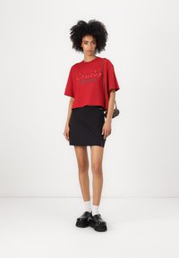 Rotes, kurzes T-Shirt mit dem Schriftzug "Coach New York", kombiniert mit einem schwarzen Minirock. Das Model trägt klobige schwarze Schuhe und weiße Socken. Minimalistischer Hintergrund.