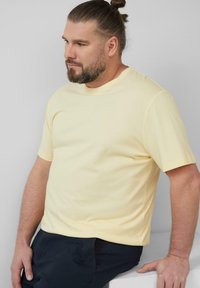 Geel T-shirt met korte mouwen en een ronde hals, gemaakt van zachte stof, met een subtiele logo geborduurd op de borst. Donkere broek.