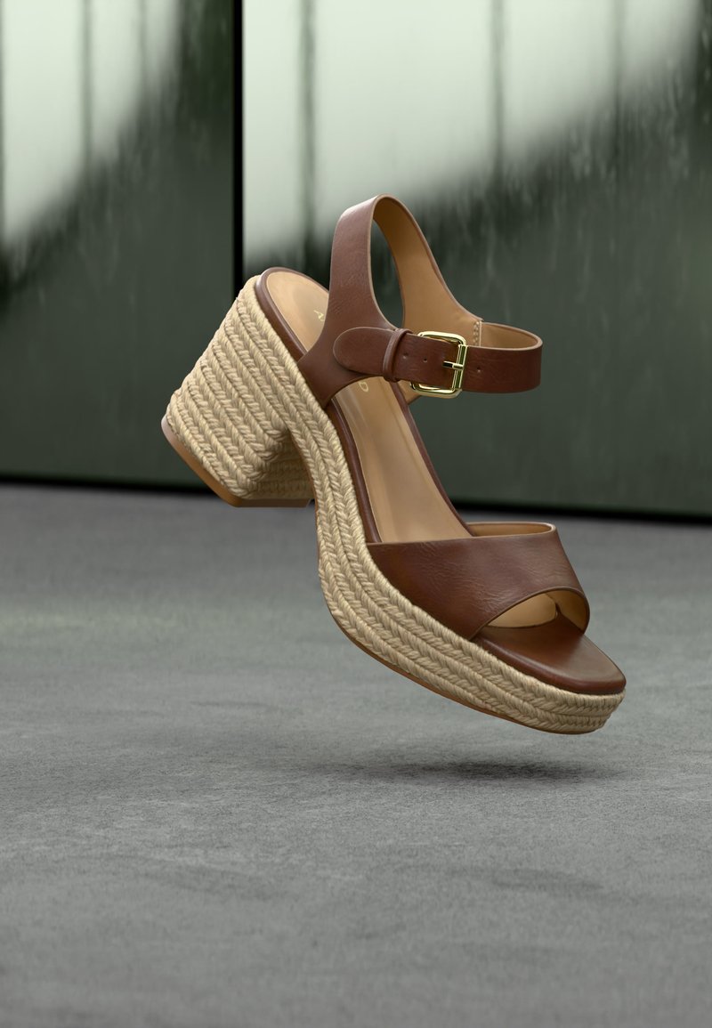 Sandale compensée en cuir marron avec bride à la cheville et boucle, dotée d'une semelle plateforme en jute tressé, suspendue au-dessus d'une surface grise.