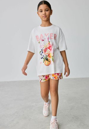 Dívka chodící v interiéru, oblečená v bílem volném tričku s květinovým vzorem, odpovídajících šortkách s motivem ovoce, bílých ponožkách a růžových teniskách.