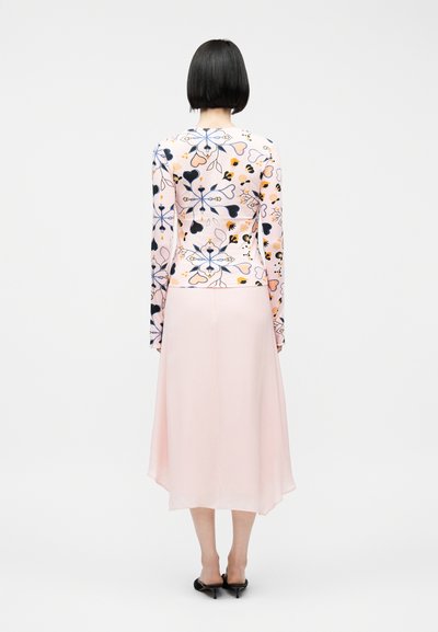 Γυναίκα με κοντά μαύρα μαλλιά, που φοράει ροζ φλοράλ μπλούζα με μακριά μανίκια και ανοιχτό ροζ midi φούστα, στέκεται με την πλάτη σε απλό φόντο.