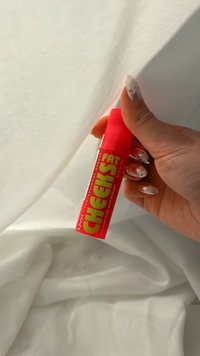 Hand houdt rode NYX Professional Makeup Fat Cheeks lippenbalsem tegen een doorschijnende witte stof achtergrond.