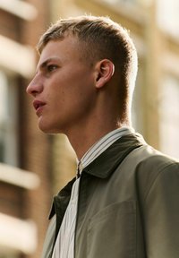 Perfil de una persona que lleva una chaqueta verde con un cuello de pana y una camisa blanca con finas rayas verticales. La luz natural resalta los rasgos.