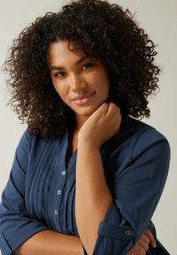 Donna con capelli ricci scuri che indossa una camicia blu navy con bottoni, toccandosi delicatamente il collo, su uno sfondo chiaro e semplice.