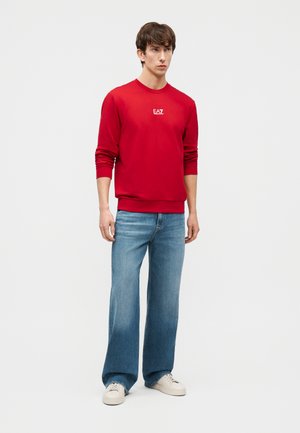 Jauns vīrietis stāv, ģērbies sarkanā EA7 garroku džempera, izbalējās, platu zilu džinsu un baltu sporta apavu apavos, uz vienkārša fona.